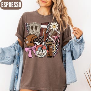 K&ouml;nnte beinhalten: Espressofarbenes T-Shirt mit einem Football-Themen-Grafikdesign. Das Design umfasst einen Helm, Fu&szlig;b&auml;lle, ein Smiley, eine G&auml;nsebl&uuml;mchen und Blitze. Das Shirt hat eine lockere Passform und hochgekrempelte &Auml;rmel.