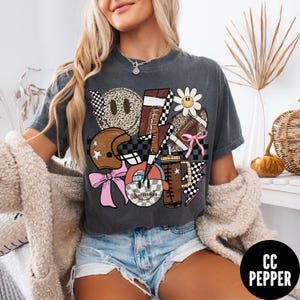 K&ouml;nnte beinhalten: Dunkelgraues T-Shirt mit einer Collage von Football-Grafiken. Das Design umfasst einen Football, einen Helm, ein Smiley, Blitze und eine G&auml;nsebl&uuml;mchen. Akzente sind rosa Schleifen und ein Schachbrettmuster.