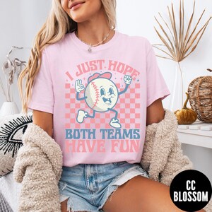 May include: Pink t-shirt with the text "I JUST HOPE BOTH TEAMS HAVE FUN" and a cartoon baseball character. The baseball is wearing a red hat and has a smiling face. The background is a pink and white checkered pattern.