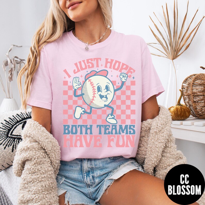 May include: Pink t-shirt with the text "I JUST HOPE BOTH TEAMS HAVE FUN" and a cartoon baseball character. The baseball is wearing a red hat and has a smiling face. The background is a pink and white checkered pattern.