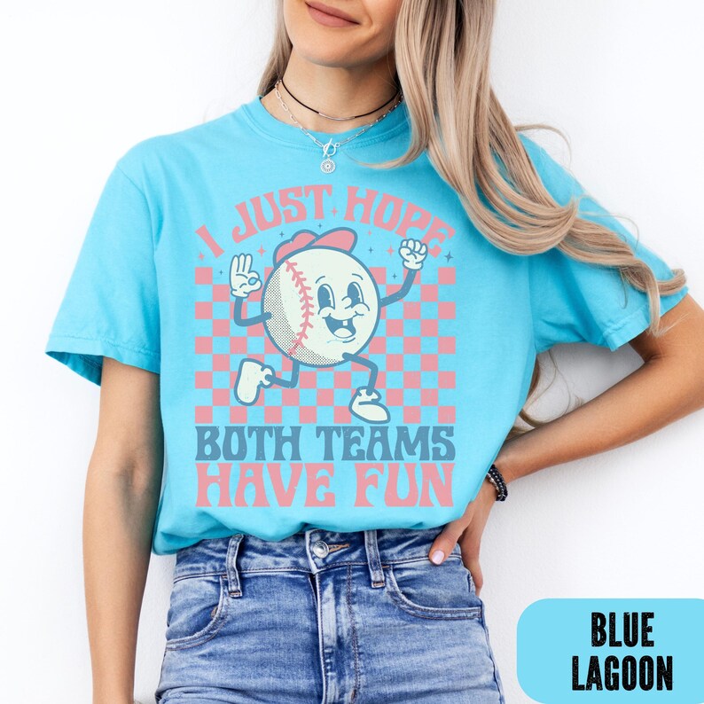 May include: A light blue t-shirt with a baseball-themed graphic. The graphic features a baseball character with the text "I JUST HOPE BOTH TEAMS HAVE FUN". The design includes pink and blue checkered squares.