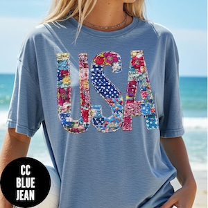 May include: A light blue t-shirt with the word "USA" in a patchwork design. The letters are filled with floral patterns and a blue and white star pattern. The shirt has short sleeves and a relaxed fit.