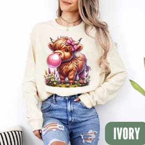 May include: Ivory-colored long-sleeve sweatshirt featuring a cartoon illustration of a Highland cow blowing a bubblegum bubble. The cow has a pink bow and is surrounded by flowers. The word "IVORY" is printed in a green rectangle.