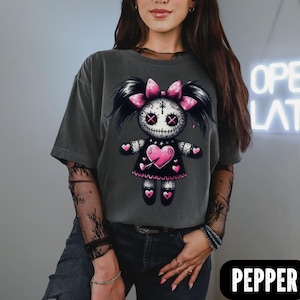 May include: A gray t-shirt with a graphic of a pink and black voodoo doll with a pink bow and hearts. The doll has a stitched-up face and is holding a pink heart with an arrow through it.