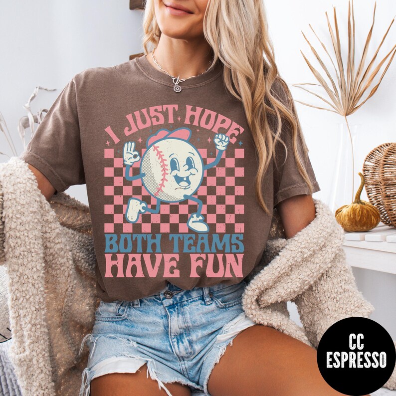 May include: Brown t-shirt with a baseball graphic. The graphic features a baseball character with the text "I JUST HOPE BOTH TEAMS HAVE FUN" in pink and blue. The background is a pink and brown checkered pattern.