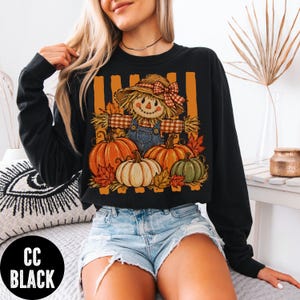 May include: A black long-sleeve sweatshirt featuring a graphic print of a scarecrow wearing a straw hat with a plaid bow, positioned above three pumpkins in orange, white, and green, accented by autumn leaves and vertical orange stripes.
