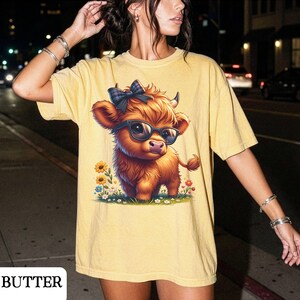 Puede incluir: Camiseta amarilla con una vaca de las Tierras Altas de dibujos animados con gafas y un lazo. La vaca est&aacute; rodeada de flores. La palabra "BUTTER" est&aacute; impresa en la parte inferior izquierda de la camiseta.