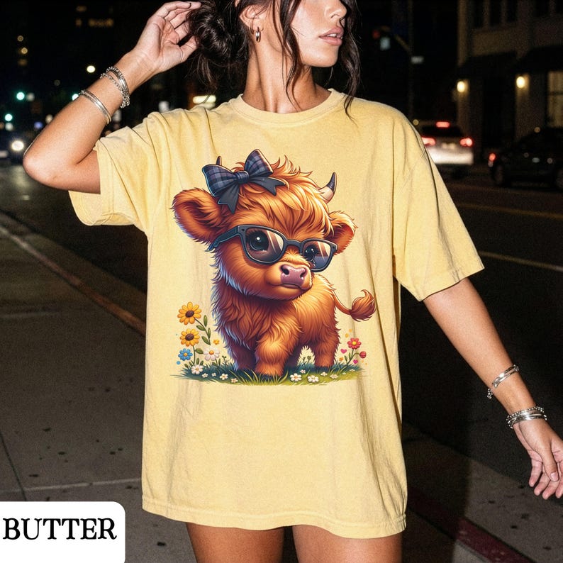 Puede incluir: Camiseta amarilla con una vaca de las Tierras Altas de dibujos animados con gafas y un lazo. La vaca est&aacute; rodeada de flores. La palabra "BUTTER" est&aacute; impresa en la parte inferior izquierda de la camiseta.