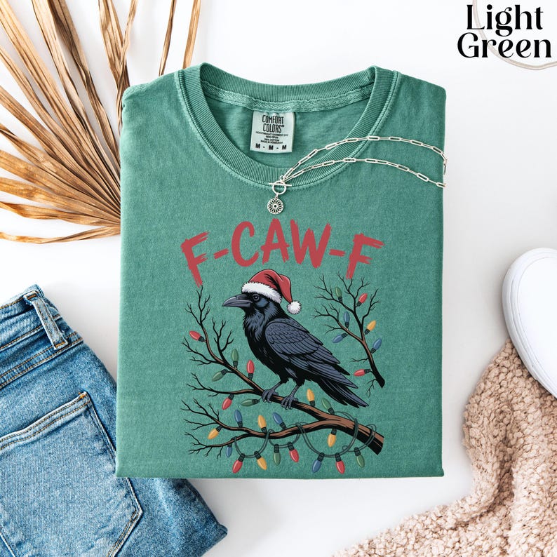 May include: A light green t-shirt with the text "F-CAW-F" in red, featuring a black crow wearing a Santa hat and perched on a branch with Christmas lights. A silver necklace is draped over the shirt.