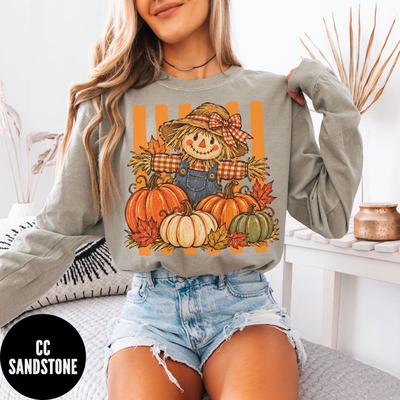 May include: A sage green long-sleeve sweatshirt featuring a graphic print of a scarecrow wearing a hat with a red plaid bow, dressed in blue overalls. The design includes orange pumpkins, a white pumpkin, a green pumpkin, and autumn leaves.