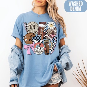 K&ouml;nnte beinhalten: Hellblaues T-Shirt mit einem Grafikdesign, das einen Football, einen Helm, Smiley-Gesichter und eine G&auml;nsebl&uuml;mchen zeigt. Das Design enth&auml;lt Schachbrettmuster, Blitze und eine rosa Schleife. Der Text "WASHED DENIM" befindet sich in einem blauen Rechteck.