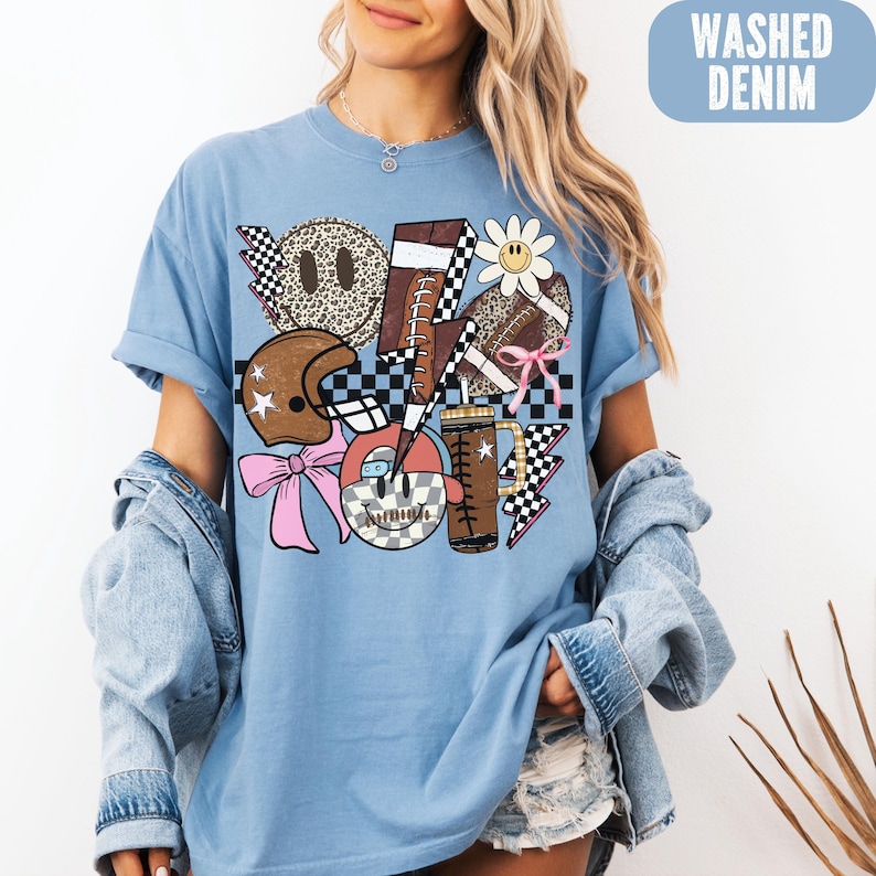 K&ouml;nnte beinhalten: Hellblaues T-Shirt mit einem Grafikdesign, das einen Football, einen Helm, Smiley-Gesichter und eine G&auml;nsebl&uuml;mchen zeigt. Das Design enth&auml;lt Schachbrettmuster, Blitze und eine rosa Schleife. Der Text "WASHED DENIM" befindet sich in einem blauen Rechteck.