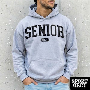 May include: A light gray hooded sweatshirt featuring the word SENIOR in large black block letters across the chest, with the number 2027 printed in a smaller black oval below. The garment includes a front kangaroo pocket and long sleeves.