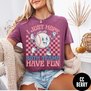 May include: A maroon t-shirt with a baseball graphic. The baseball character has a smiling face and is wearing a baseball cap. The text on the shirt reads "I JUST HOPE BOTH TEAMS HAVE FUN".