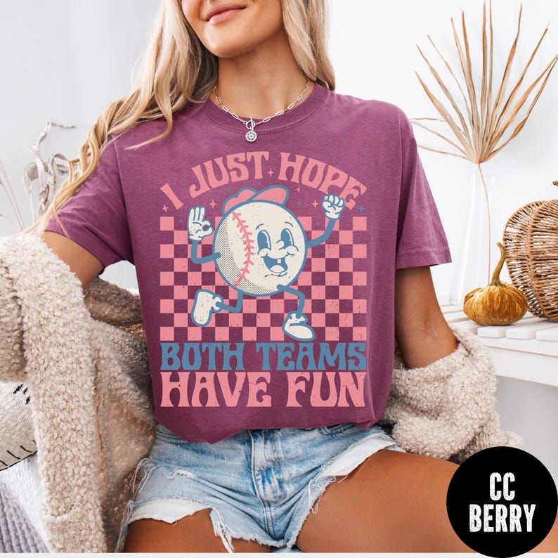 May include: A maroon t-shirt with a baseball graphic. The baseball character has a smiling face and is wearing a baseball cap. The text on the shirt reads "I JUST HOPE BOTH TEAMS HAVE FUN".