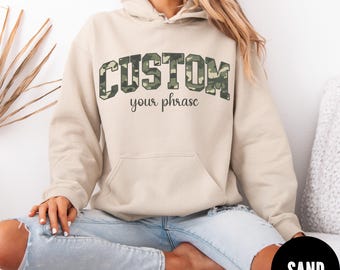 May include: A sand-colored hoodie with a front pocket and drawstring hood. The word "CUSTOM" is printed across the chest in a camouflage pattern, with "your phrase" printed below in a script font. The hoodie is worn with blue jeans.