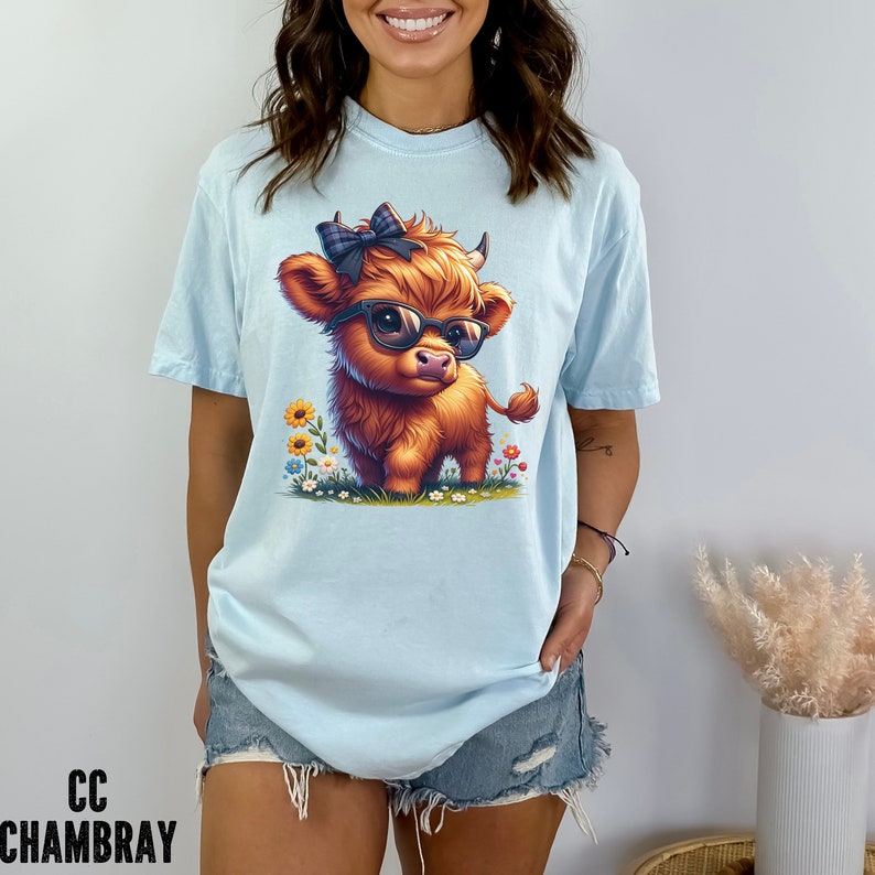Puede incluir: Camiseta azul claro con una vaca de las Tierras Altas de dibujos animados con gafas de sol y un lazo a cuadros. La vaca est&aacute; rodeada de flores coloridas. El texto "CC CHAMBRAY" est&aacute; impreso en la parte inferior izquierda.