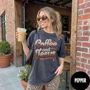 May include: Dark gray t-shirt with the words "Coffee and Jesus" in a brown and cream-colored design. The shirt is worn by a person with sunglasses, holding an iced coffee. The background includes a brick wall and a coffee shop.