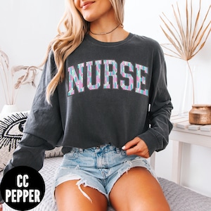 May include: A dark gray long-sleeved cropped sweatshirt with the word "NURSE" printed in pink, blue, and white letters with a patterned design. The sweatshirt is worn by a person wearing denim shorts.