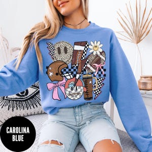 May include: Carolina blue sweatshirt with a graphic design featuring a football, lightning bolts, a daisy, a football helmet, and a smiley face. The design incorporates checkered patterns and a pink bow. The sweatshirt has a crew neck.