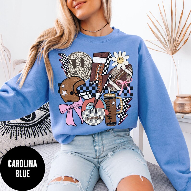 May include: Carolina blue sweatshirt with a graphic design featuring a football, lightning bolts, a daisy, a football helmet, and a smiley face. The design incorporates checkered patterns and a pink bow. The sweatshirt has a crew neck.