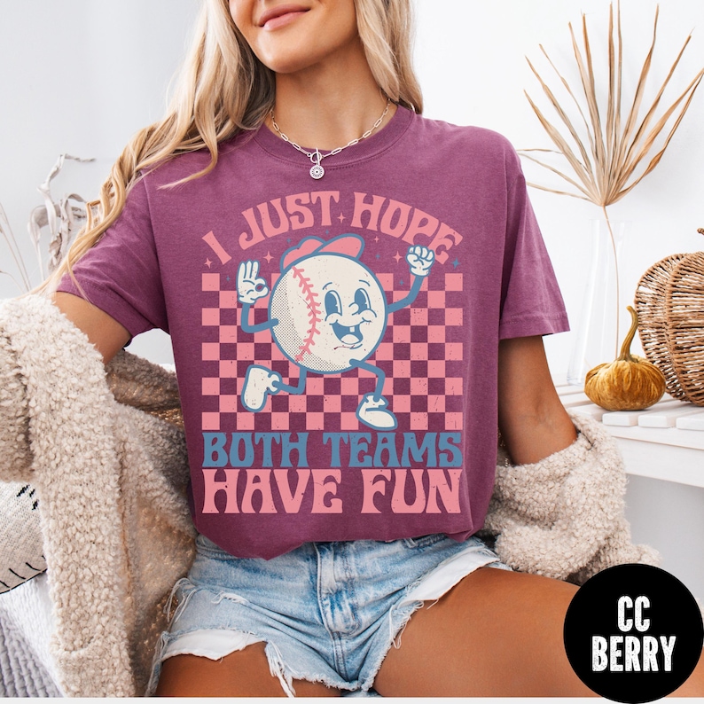 May include: A maroon t-shirt with a baseball graphic. The baseball character has a smiling face and is wearing a baseball cap. The text on the shirt reads "I JUST HOPE BOTH TEAMS HAVE FUN".