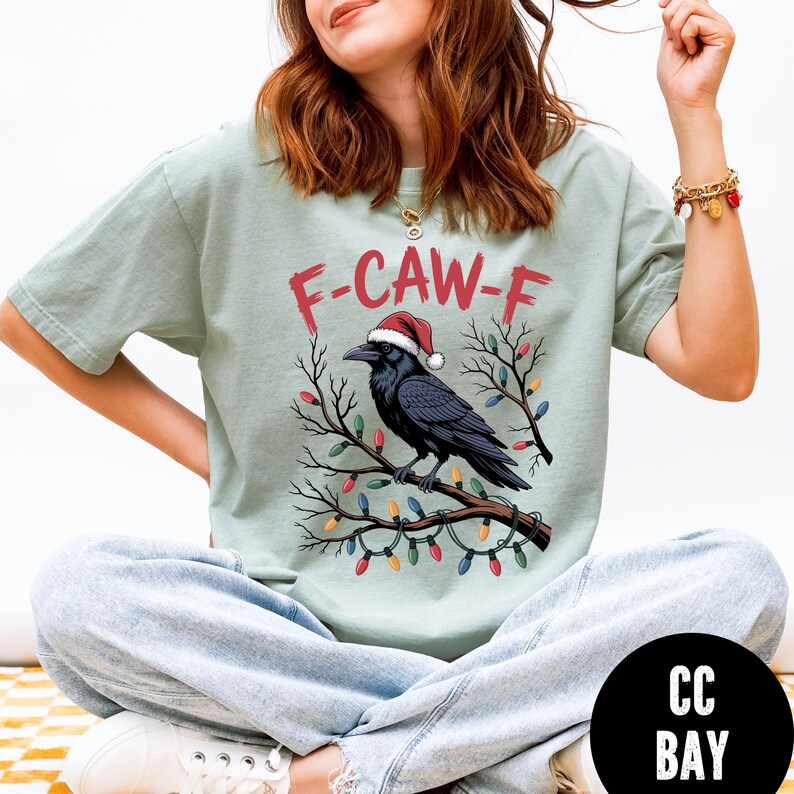 May include: Sage green t-shirt featuring a black crow wearing a Santa hat, perched on a branch decorated with Christmas lights. The text "F-CAW-F" is above the crow. A festive holiday-themed graphic tee.