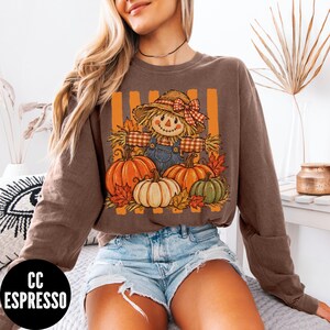 May include: A brown long-sleeve sweatshirt featuring a graphic print of a scarecrow wearing a straw hat with a red plaid bow, surrounded by orange, white, and green pumpkins and autumn leaves against a background of vertical orange stripes.