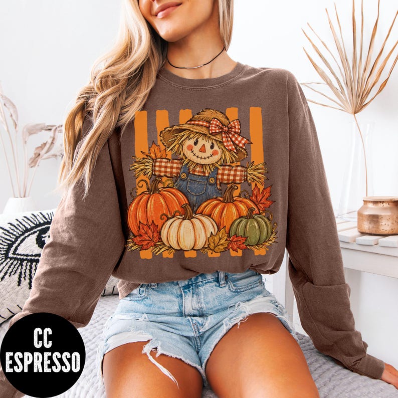 May include: A brown long-sleeve sweatshirt featuring a graphic print of a scarecrow wearing a straw hat with a red plaid bow, surrounded by orange, white, and green pumpkins and autumn leaves against a background of vertical orange stripes.