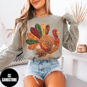 May include: A long-sleeved, olive green shirt featuring a colorful turkey design. The turkey is made of various patterned fabrics in shades of red, orange, yellow, and brown. The shirt has a crew neck and long sleeves, perfect for fall.