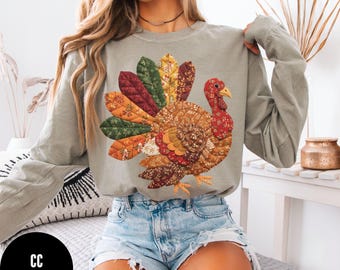 May include: An olive green long-sleeved top with a detailed turkey design. The turkey is crafted from patchwork fabrics in autumnal colours: red, orange, yellow, and brown. The top has a round neck and long sleeves, suitable for cooler weather.