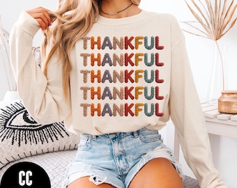 May include: Cream-colored long-sleeve sweatshirt with the word "THANKFUL" in colorful, textured letters. The text is stacked vertically. The model is wearing denim shorts. The sweatshirt is from the brand "CC IVORY".