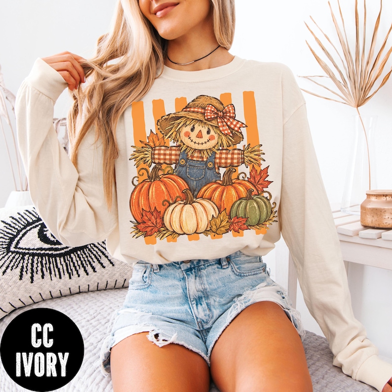 May include: An ivory long-sleeve sweatshirt featuring a graphic print of a scarecrow wearing a straw hat with a red plaid bow, surrounded by orange and green pumpkins and autumn leaves against a background of vertical orange stripes.