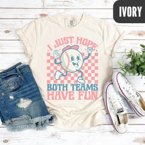 May include: Ivory-colored t-shirt with a baseball graphic and the text "I JUST HOPE BOTH TEAMS HAVE FUN." The baseball character is wearing a hat and has arms and legs. The background is a pink and white checkered pattern. The shirt is paired with denim shorts and white sneakers.