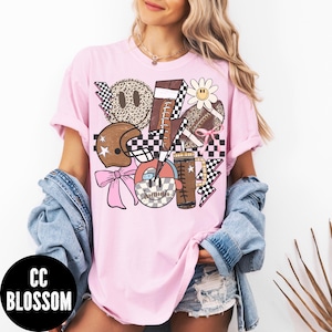 K&ouml;nnte beinhalten: Hellrosa T-Shirt mit einem Grafikdesign mit Footballs, einem Helm, einer G&auml;nsebl&uuml;mchen und einem Kaffeebecher. Das Design enth&auml;lt Schachbrettmuster, Blitze und ein Smiley-Gesicht. Der Text "CC BLOSSOM" befindet sich in einem schwarzen Kreis.