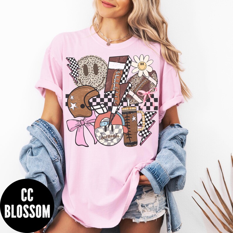 K&ouml;nnte beinhalten: Hellrosa T-Shirt mit einem Grafikdesign mit Footballs, einem Helm, einer G&auml;nsebl&uuml;mchen und einem Kaffeebecher. Das Design enth&auml;lt Schachbrettmuster, Blitze und ein Smiley-Gesicht. Der Text "CC BLOSSOM" befindet sich in einem schwarzen Kreis.