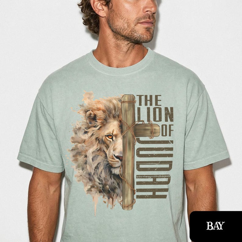 K&ouml;nnte beinhalten: Hellgr&uuml;nes T-Shirt mit einer detaillierten Illustration eines L&ouml;wengesichts und eines Holzkreuzes. Der Text lautet "THE LION OF JUDAH" in einer vertikalen Schriftart. Das T-Shirt ist einfarbig und hat kurze &Auml;rmel.
