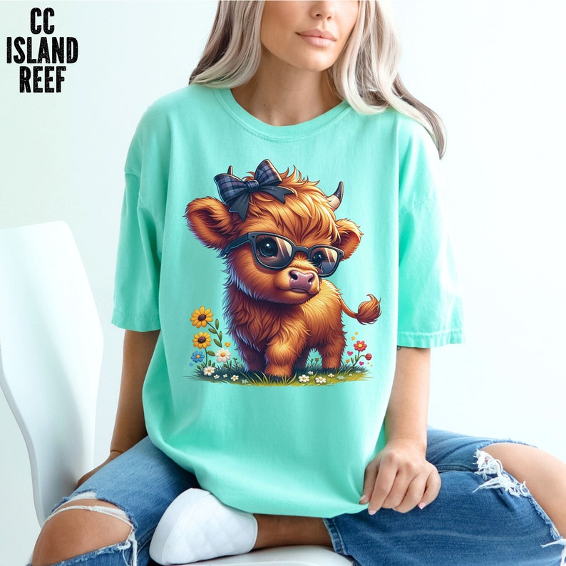 Puede incluir: Camiseta verde azulado claro con una ilustraci&oacute;n de dibujos animados de una vaca de las Tierras Altas con gafas y un lazo a cuadros. La vaca marr&oacute;n tiene cuernos peque&ntilde;os y est&aacute; rodeada de flores. El texto "CC ISLAND REEF" est&aacute; en la esquina superior izquierda.
