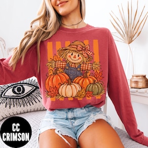 May include: A long-sleeve crewneck sweatshirt in a muted red color featuring a graphic print of a scarecrow wearing a straw hat with a red plaid bow, dressed in blue overalls, surrounded by orange, white, and green pumpkins and autumn leaves.