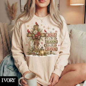 May include: A cream-colored long-sleeve sweatshirt featuring a graphic of a wooden cross adorned with red cardinals, poinsettias, and a plaid bow. Below the cross is a lantern and stacked wooden signs reading Jesus is the reason for the season.