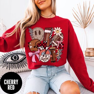 May include: Cherry red sweatshirt with a graphic design featuring a football, helmet, smiley faces, and lightning bolts. The design incorporates checkered patterns, a daisy, and a pink bow. The text "CHERRY RED" is in a black circle.