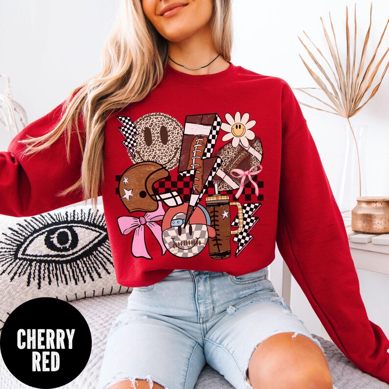 May include: Cherry red sweatshirt with a graphic design featuring a football, helmet, smiley faces, and lightning bolts. The design incorporates checkered patterns, a daisy, and a pink bow. The text "CHERRY RED" is in a black circle.