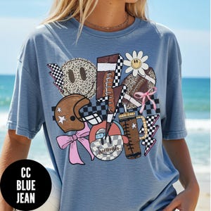 K&ouml;nnte beinhalten: Hellblaues Jeans-T-Shirt mit einem Grafikdesign mit Football, Helm, Smiley-Gesichtern und einer G&auml;nsebl&uuml;mchen. Das Design enth&auml;lt Schachflaggen, Blitze und eine rosa Schleife. Der Text "CC BLUE JEAN" befindet sich in einem schwarzen Kreis.