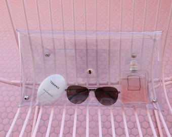 May include: A clear plastic clutch bag with a zipper closure. The bag contains a white tube of La Creme Main hand cream by Chanel, a pair of brown sunglasses, and a clear bottle of perfume.