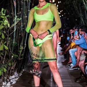 May include: A model walks the runway wearing a neon green mesh dress with a hood. The dress is sheer and shows off the model's figure. The model is also wearing black fishnet stockings and green heels. The model is carrying a clear plastic bag with the words "Fashion Store" printed on it.