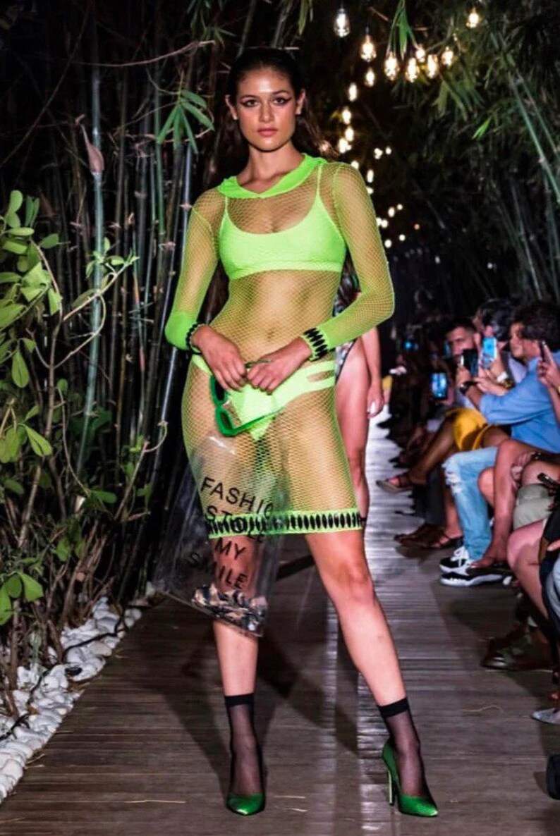 May include: A model walks the runway wearing a neon green mesh dress with a hood. The dress is sheer and shows off the model's figure. The model is also wearing black fishnet stockings and green heels. The model is carrying a clear plastic bag with the words "Fashion Store" printed on it.