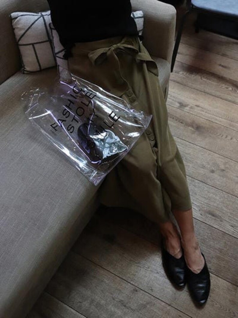 May include: A person wearing a green skirt and black shoes is holding a clear plastic bag with the text "Fashion Hole Store" printed on it.