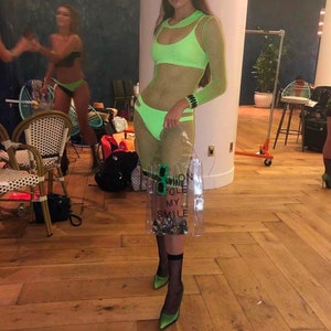 May include: A woman wearing a green mesh dress with a green bikini top and matching bikini bottoms. She is holding a clear plastic bag with the text "Fashion Toile My Smile" printed on it. She is wearing green heels and black fishnet stockings.
