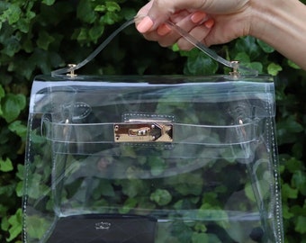 May include: A clear, rectangular handbag with a gold-colored clasp and handle attachments. The bag has a transparent design, allowing visibility of the contents inside, including a black wallet. The handbag is held by a person.