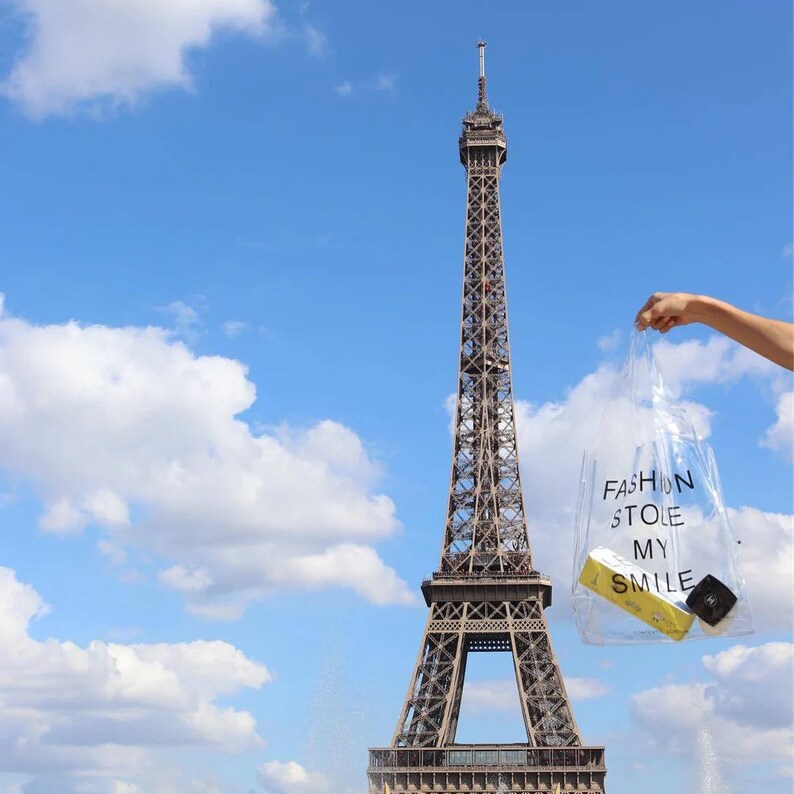 May include: A clear plastic bag with the text "FASHION STOLE MY SMILE" printed on it. The bag is being held in front of the Eiffel Tower in Paris, France.